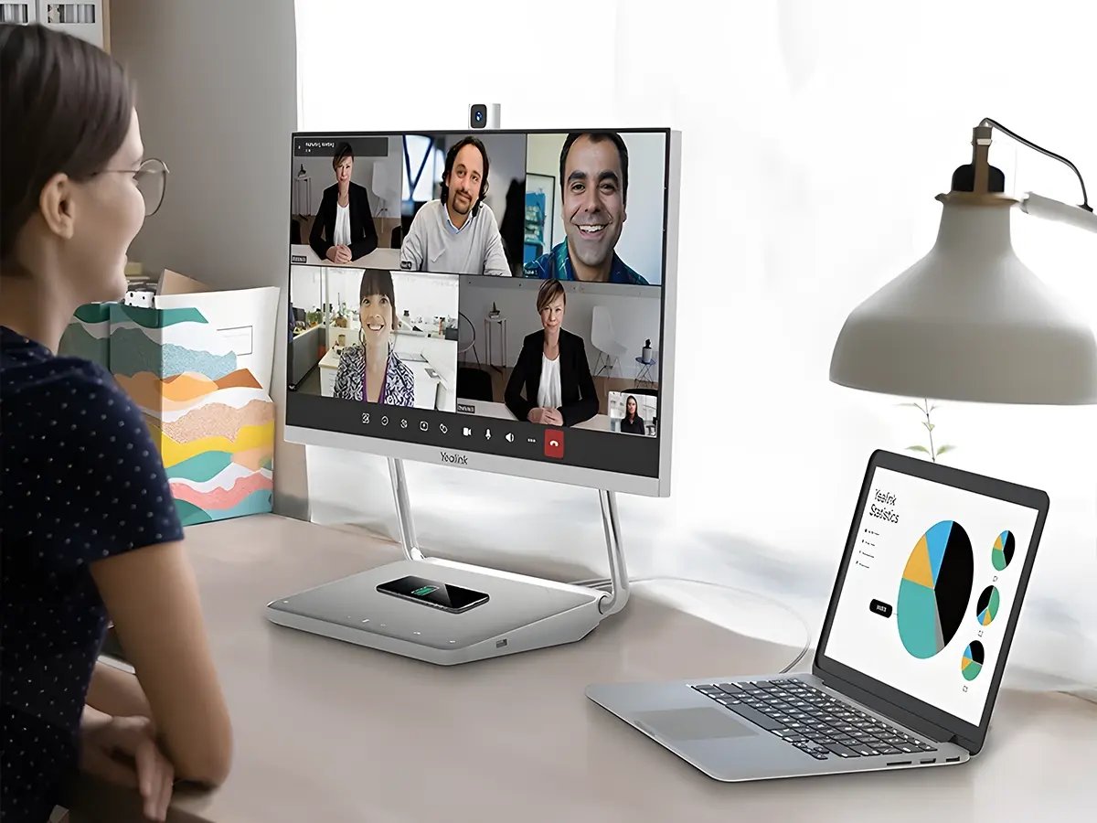 Person seated at a desk participating in a video call displayed on a large monitor in a home office.