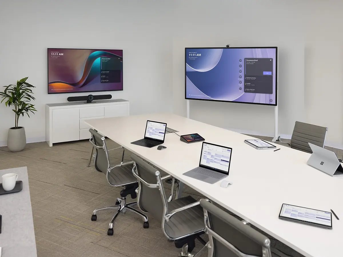 Modern meeting room with a long table, chairs and two large wall‑mounted displays showing presentation content.