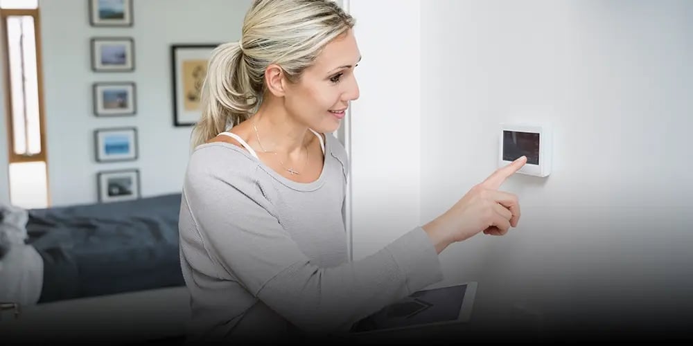A woman adjusts a smart thermostat on a wall, smiling and holding a tablet. The room is modern and bright, conveying a sense of comfort and technology.