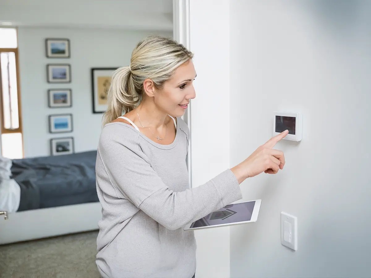 A woman adjusts a smart thermostat on a wall, smiling and holding a tablet. The room is modern and bright, conveying a sense of comfort and technology.