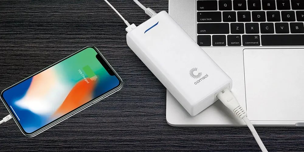 A smartphone and a MacBook on a dark wood desk, both connected to a white power bank with cables. The phone shows a charging display.