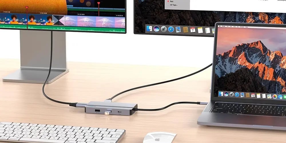 A laptop connects to two monitors via a USB hub on a wooden desk. A keyboard and mouse are nearby, displaying a neat workspace setup.