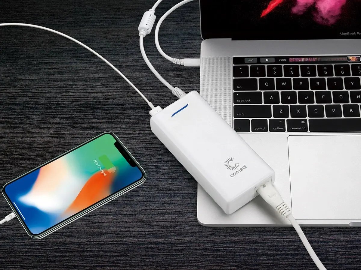 A smartphone and a MacBook on a dark wood desk, both connected to a white power bank with cables. The phone shows a charging display.
