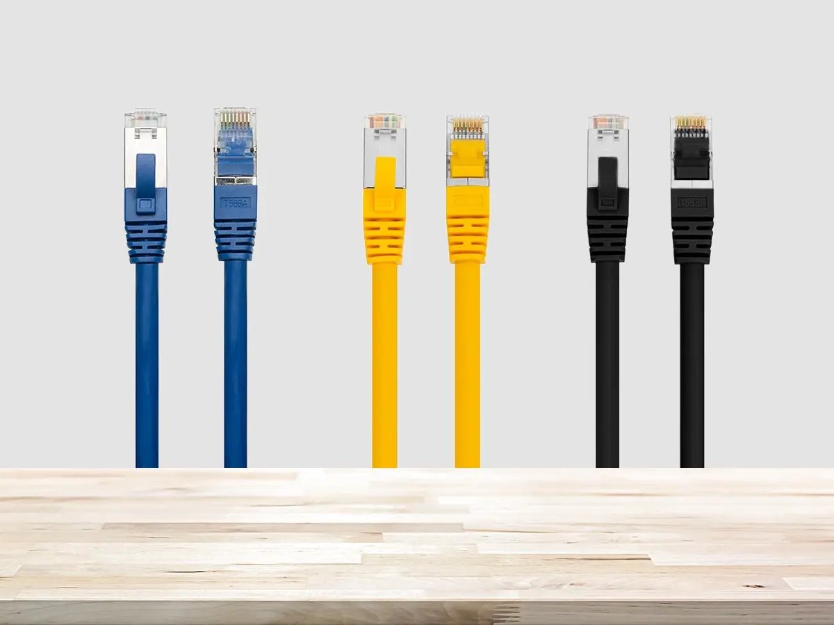 Three Ethernet cables in blue, yellow, and black are aligned vertically against a light gray background, above a wooden surface.