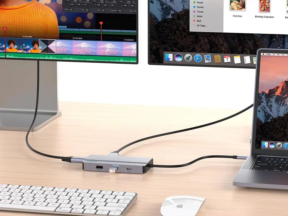 A laptop connects to two monitors via a USB hub on a wooden desk. A keyboard and mouse are nearby, displaying a neat workspace setup.