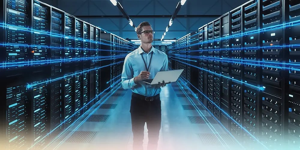 A man with glasses holds a tablet in a modern server room, surrounded by rows of servers with glowing blue lines, conveying a high-tech ambiance.