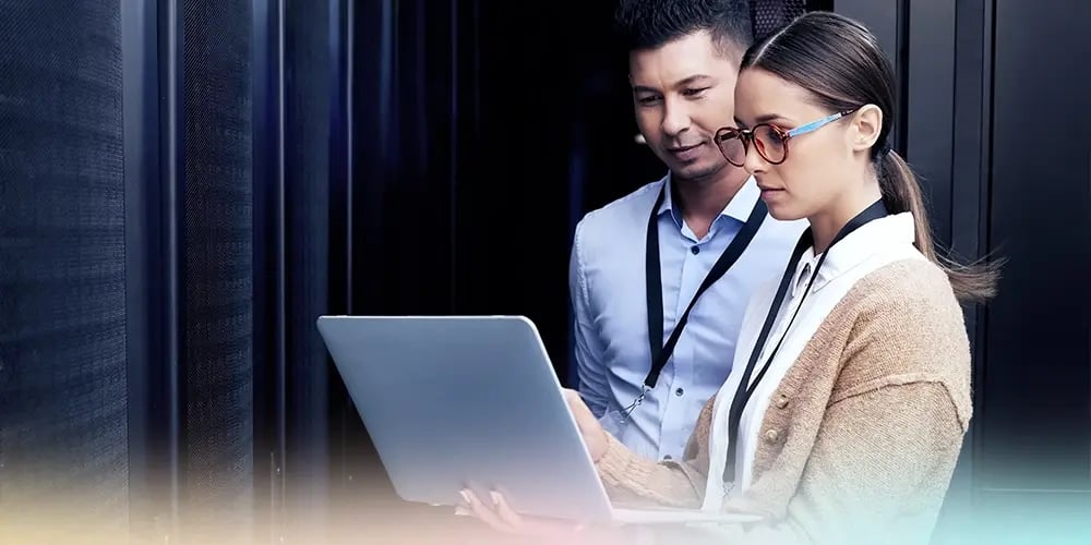 Two professionals, one male and one female, stand in a server room reviewing data on a laptop. They appear focused and collaborative, dressed in business attire.