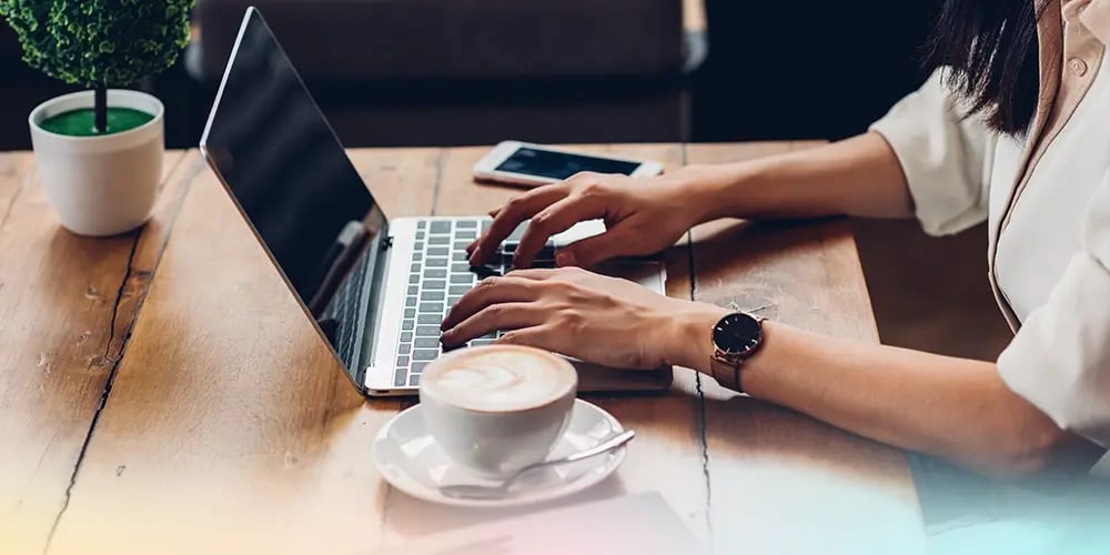 A person types on a laptop at a wooden table, with a cappuccino and smartphone nearby. The scene is bright and focused, suggesting productivity.