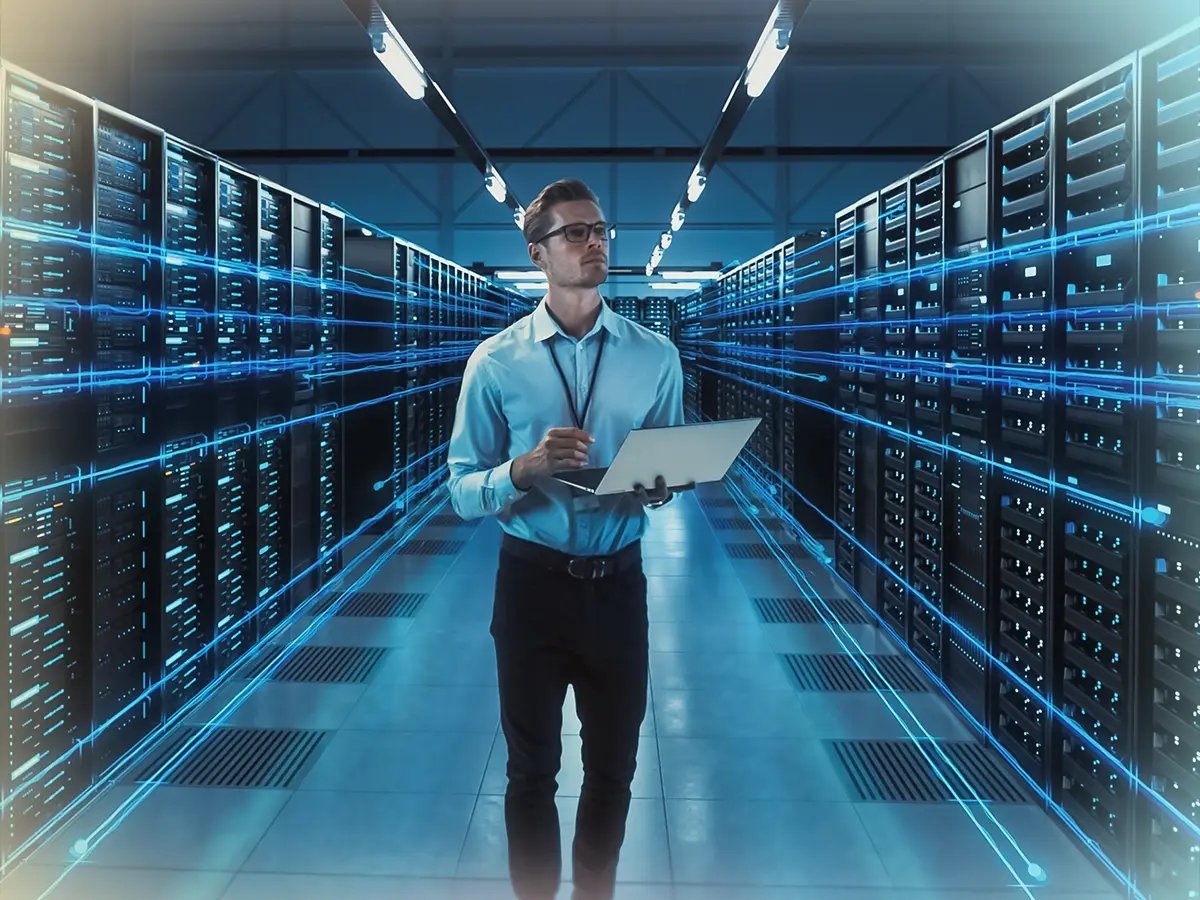 A man with glasses holds a tablet in a modern server room, surrounded by rows of servers with glowing blue lines, conveying a high-tech ambiance.