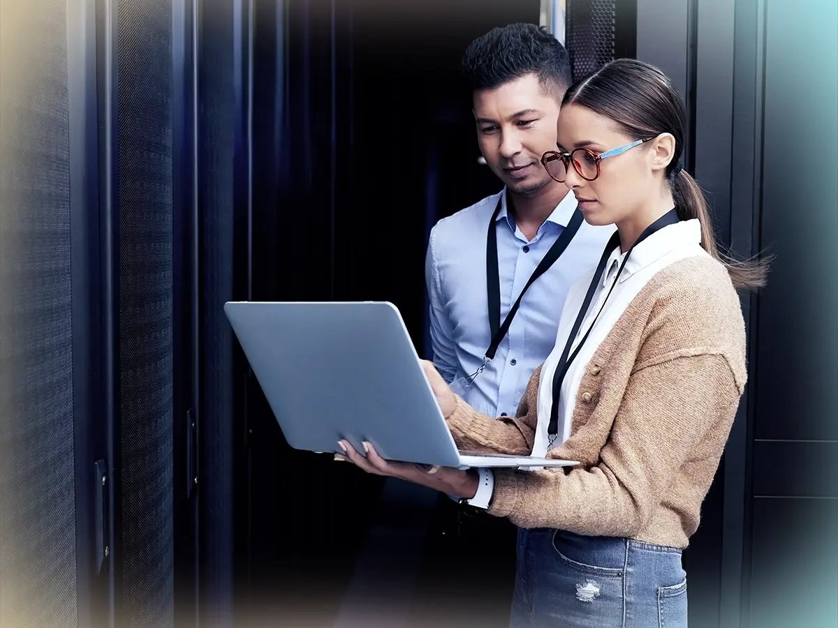 Two professionals, one male and one female, stand in a server room reviewing data on a laptop. They appear focused and collaborative, dressed in business attire.