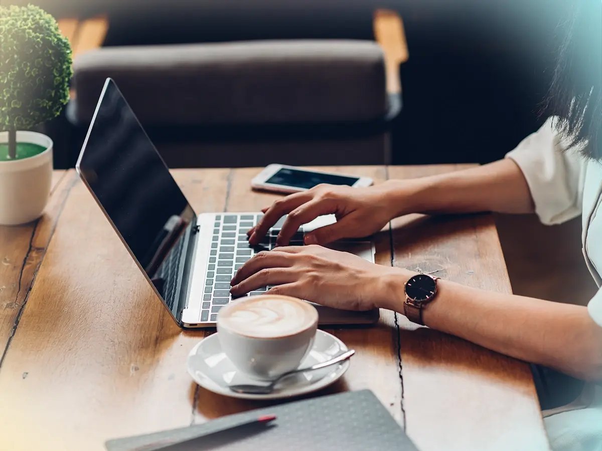 A person types on a laptop at a wooden table, with a cappuccino and smartphone nearby. The scene is bright and focused, suggesting productivity.