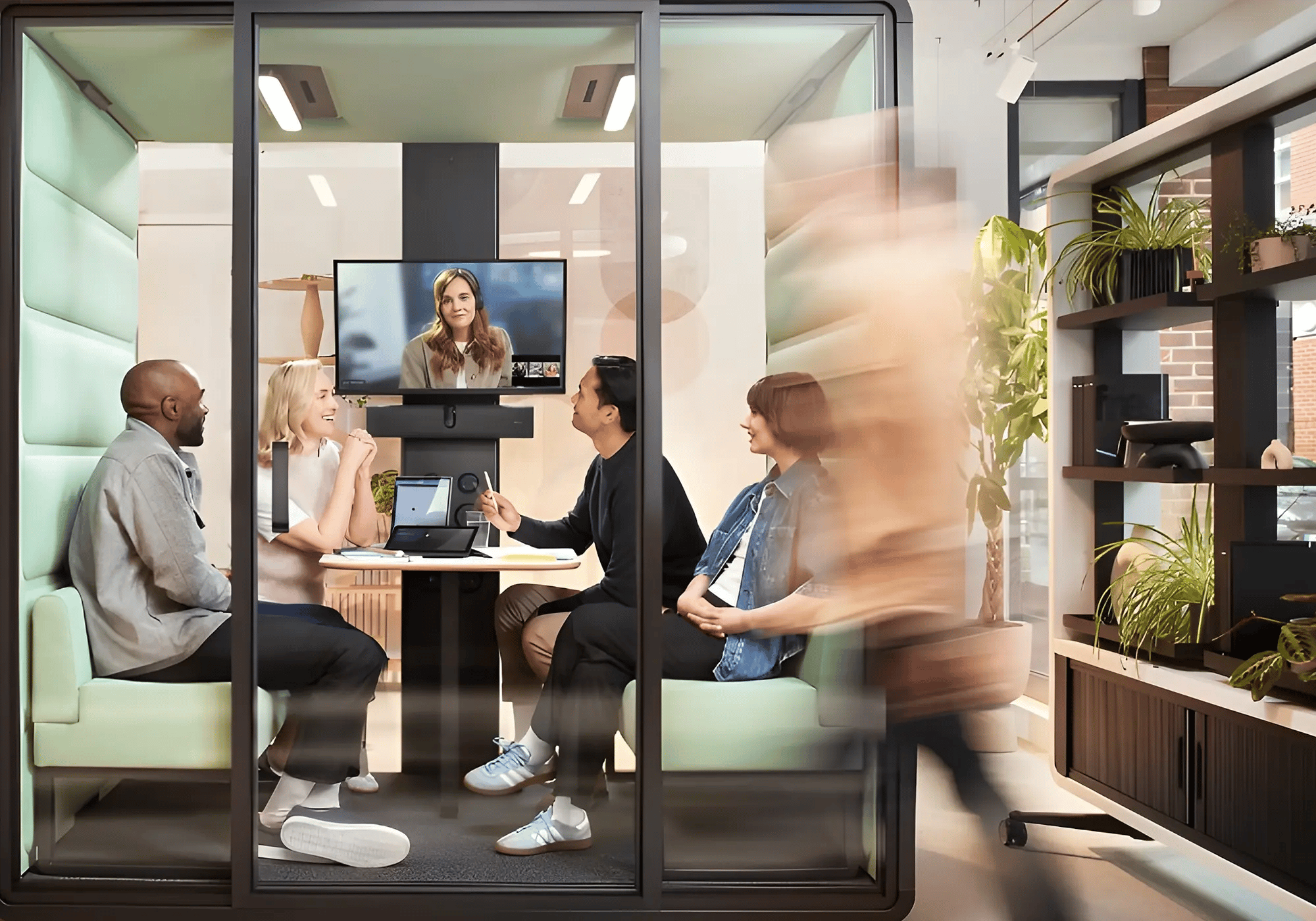 A group of four people sit in a modern meeting pod, engaging in a video call with a woman on a screen. The space is bright, with a blurred figure walking by, creating a lively atmosphere.