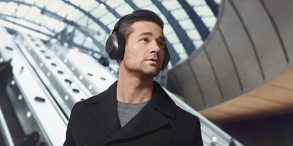A man with dark hair and a black coat listens to headphones in a modern, glass-structured building. He looks contemplative while standing near escalators.