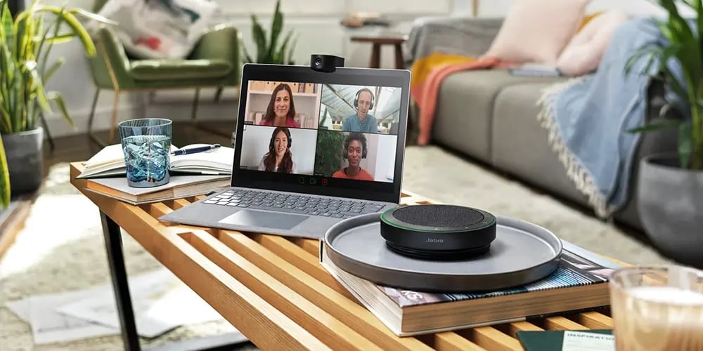 A cozy living room features a laptop on a wooden coffee table, displaying a video call with four smiling participants. Books, a glass of water, and a speaker sit nearby, exuding a warm, collaborative atmosphere.