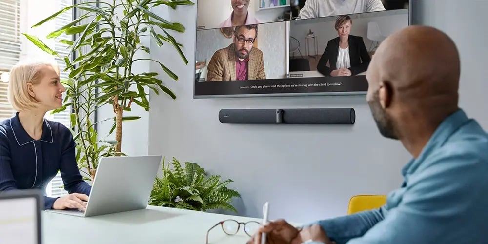 Two colleagues in an office setting participate in a video conference on a wall screen. The setting is modern, with plants and a relaxed atmosphere.