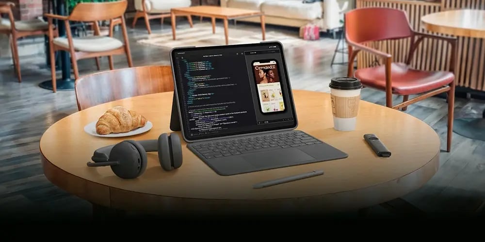 A remote work setup in a café environment featuring Logitech devices, including wireless headphones and a stylus placed beside a tablet with a keyboard displaying code and a mobile app preview. A coffee cup and croissant sit on the table, illustrating a flexible, on-the-go workspace designed for working remotely.