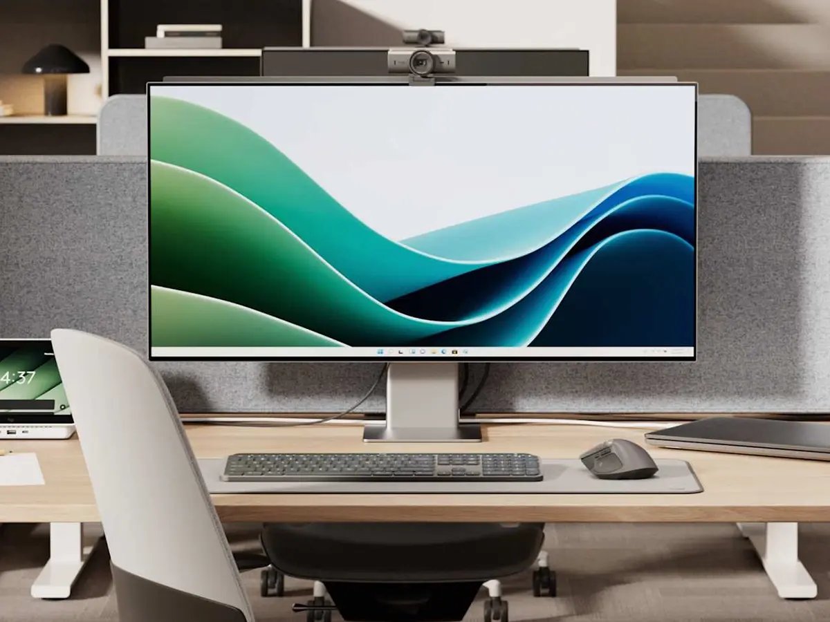 A modern personal workspace featuring Logitech devices, including a widescreen monitor with a webcam mounted on top, a wireless keyboard and mouse, and a headset resting on the desk. The setup sits on a light wood desk with a fabric privacy panel, tablet, and notebook, creating a clean, professional home or office work environment.