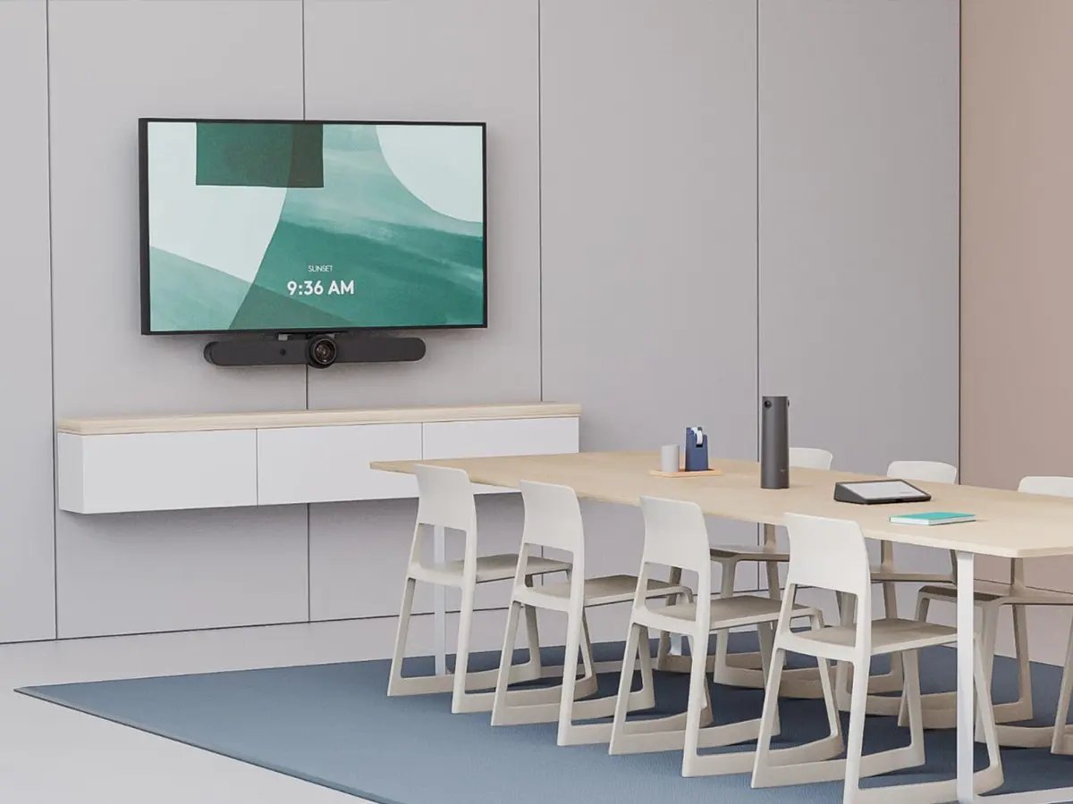 A modern conference room equipped with Logitech meeting room products, featuring a wall-mounted display with a video bar beneath it, a long table with multiple chairs, and a tabletop conference microphone. A whiteboard with notes and a clean, minimalist interior highlight a professional collaboration space designed for video meetings.