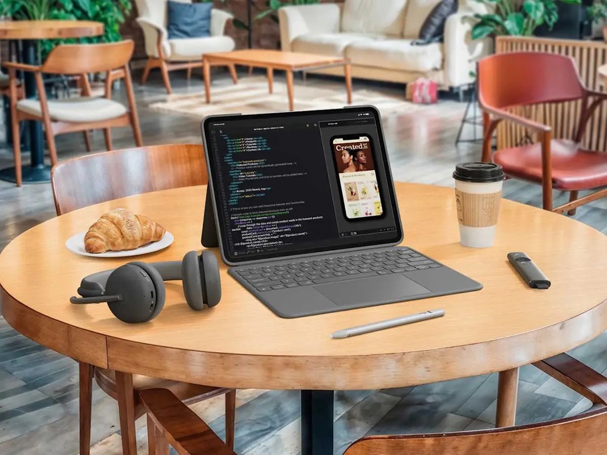 A remote work setup in a café environment featuring Logitech devices, including wireless headphones and a stylus placed beside a tablet with a keyboard displaying code and a mobile app preview. A coffee cup and croissant sit on the table, illustrating a flexible, on-the-go workspace designed for working remotely.