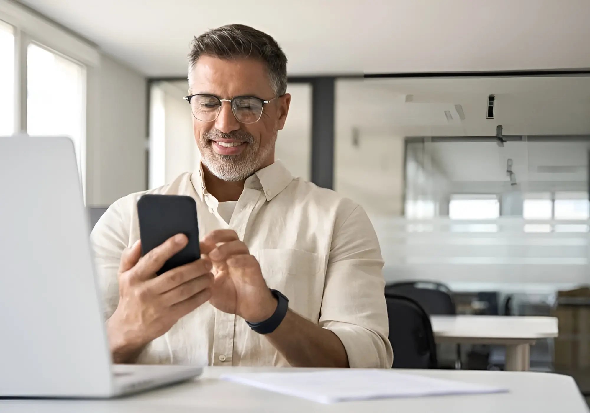 Smiling man with glasses uses a smartphone while sitting at a desk with a laptop. Bright office setting, conveying a positive and focused atmosphere.