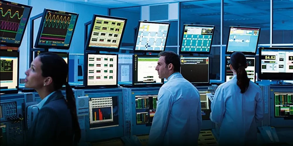 Three people in lab coats are focused on multiple monitors displaying complex data, in a brightly lit control room, conveying focus and collaboration.