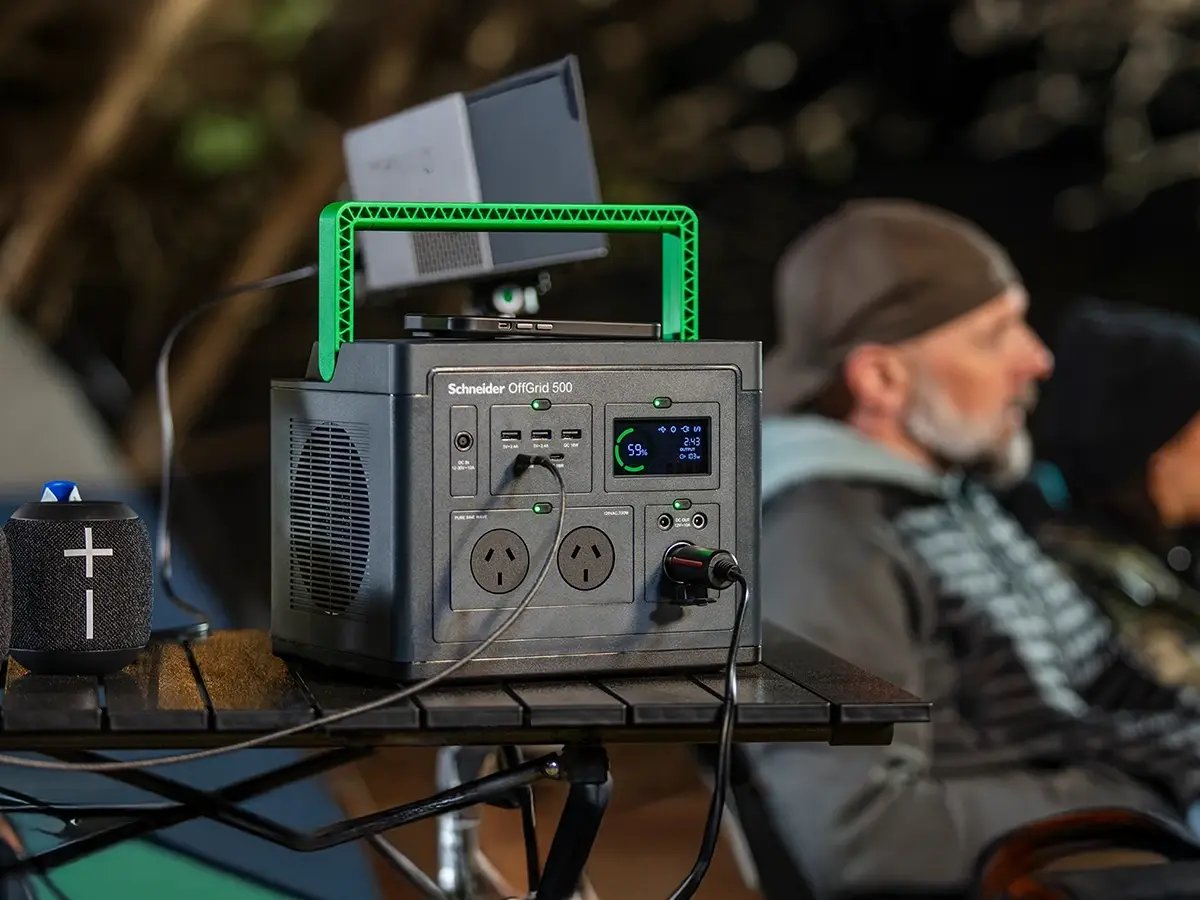 A portable power station with a green handle sits on a table, charging devices. In the background, two people sit, blurred, wearing outdoor gear.