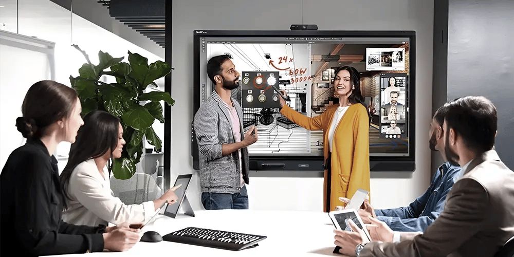 A group of six people sits in a modern conference room, engaging with a large interactive screen. Two are presenting ideas enthusiastically, while others listen attentively, holding tablets. The atmosphere is collaborative and tech-driven.