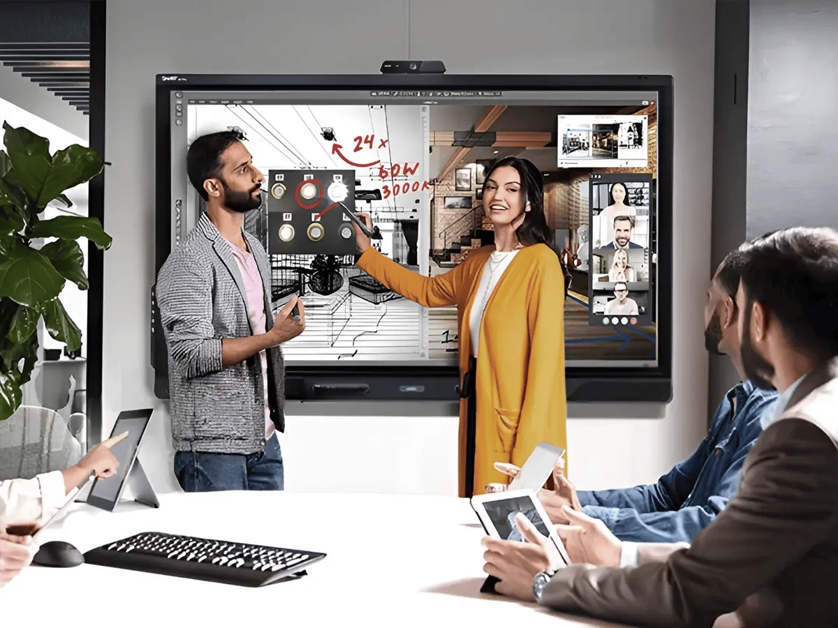 A group of six people sits in a modern conference room, engaging with a large interactive screen. Two are presenting ideas enthusiastically, while others listen attentively, holding tablets. The atmosphere is collaborative and tech-driven.