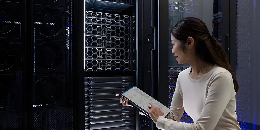 A woman examines a server rack while holding a tablet. The room is dimly lit, highlighting the sleek, modern technology. The tone is professional and focused.