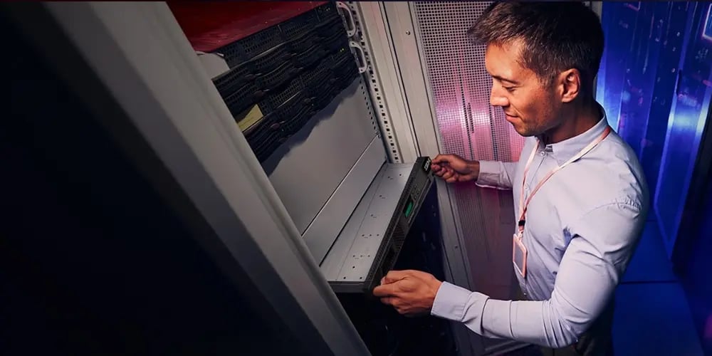 Man in a white shirt working in a server room, sliding a server into a rack. The environment is professional and technical, with a focused expression.