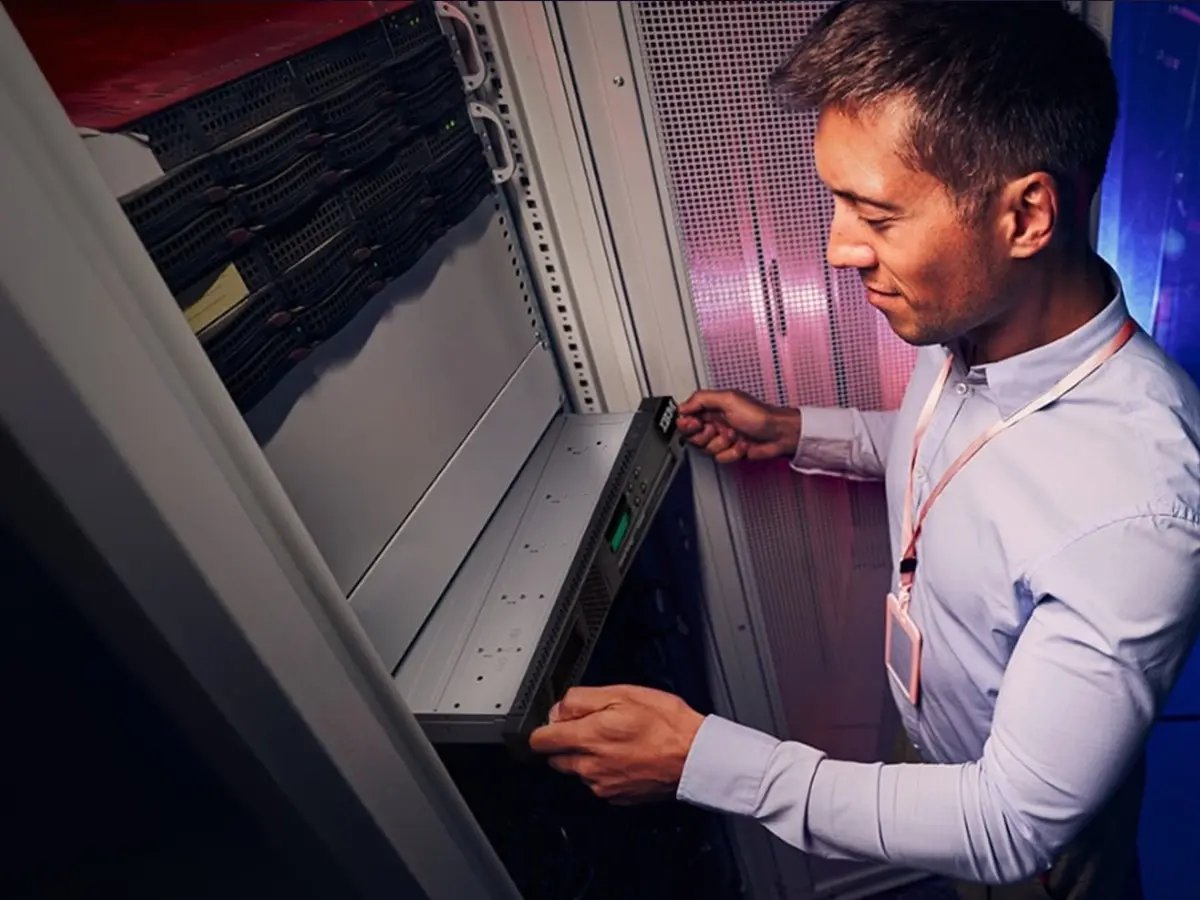 Man in a white shirt working in a server room, sliding a server into a rack. The environment is professional and technical, with a focused expression.