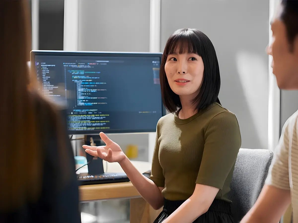 A woman in a green shirt gestures while discussing something with two others. Behind her is a computer screen displaying code. The atmosphere is collaborative.