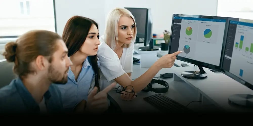 Three people are focused on computer screens displaying graphs and charts in an office. They show interest and concentration, pointing at the data.