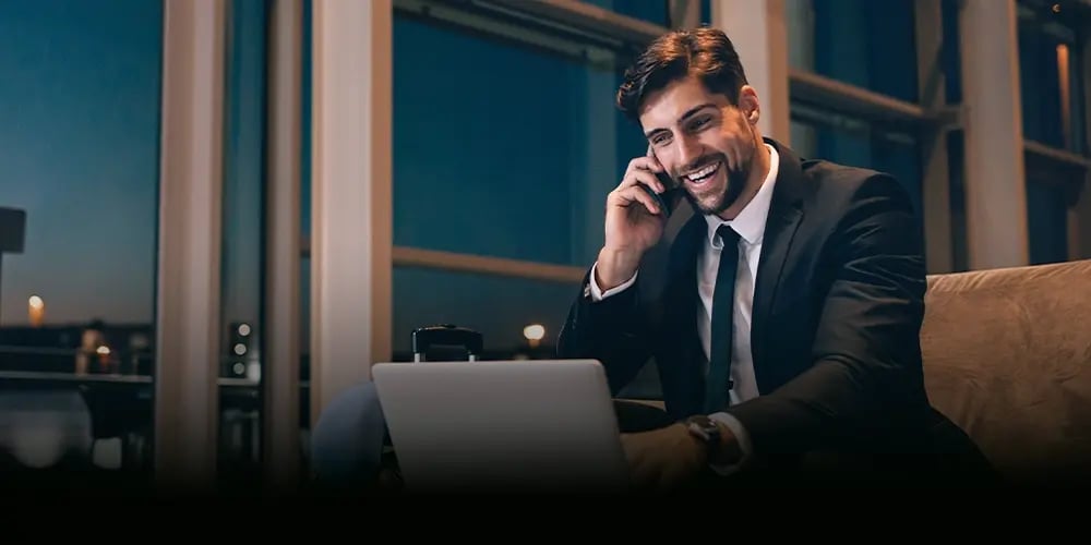 A man in a suit talks on a phone, smiling, while using a laptop. He's sitting in a modern office with large windows. It’s evening, and the mood is positive.