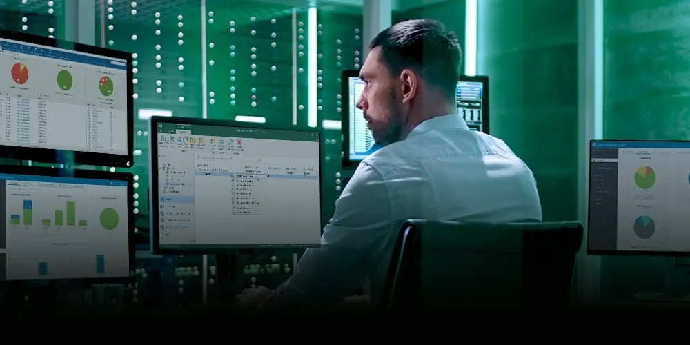 A man in a white shirt works in a dimly-lit data center. He is seated at a desk with multiple screens displaying charts and graphs. The ambience is focused and analytical.