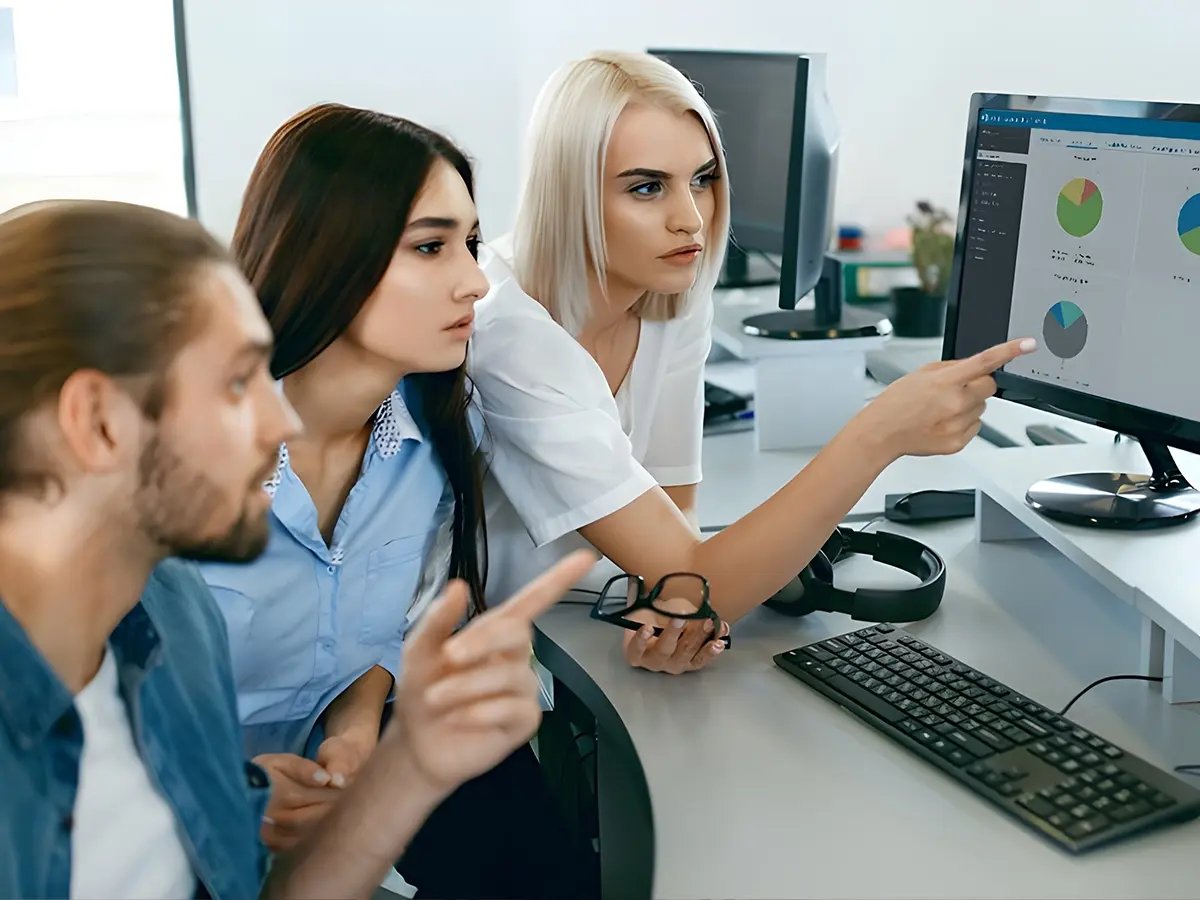 Three people are focused on computer screens displaying graphs and charts in an office. They show interest and concentration, pointing at the data.