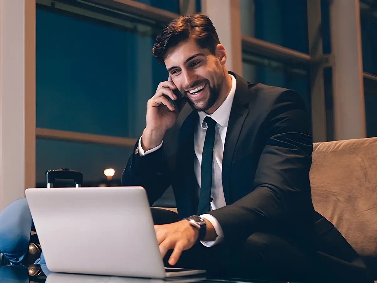 A man in a suit talks on a phone, smiling, while using a laptop. He's sitting in a modern office with large windows. It’s evening, and the mood is positive.