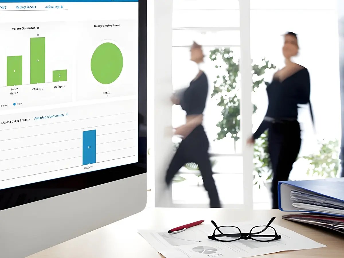 A computer screen displays bar charts and graphs in an office setting. In the blurred background, three people walk by, creating a dynamic and busy atmosphere.