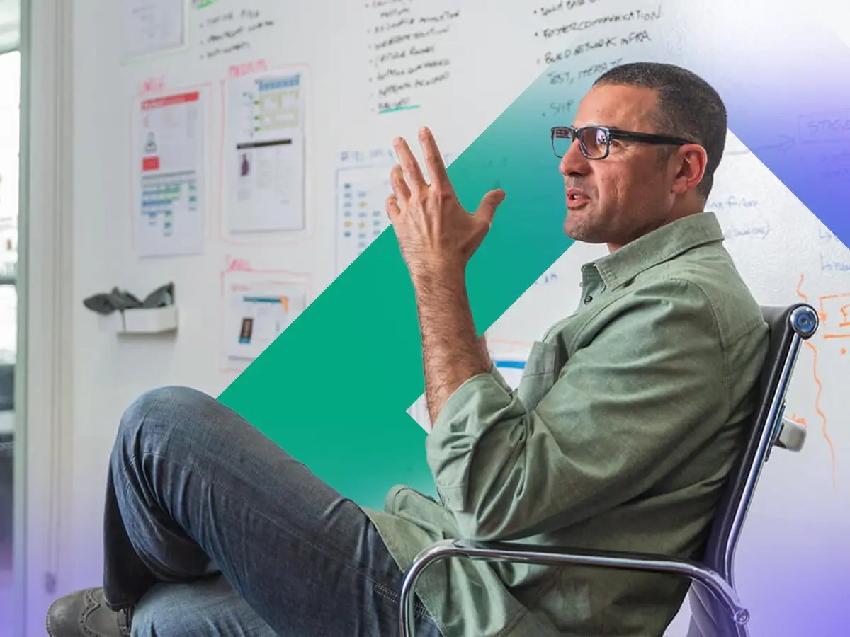 Person sitting on a chair in front of a whiteboard covered with notes and diagrams, gesturing with one hand.