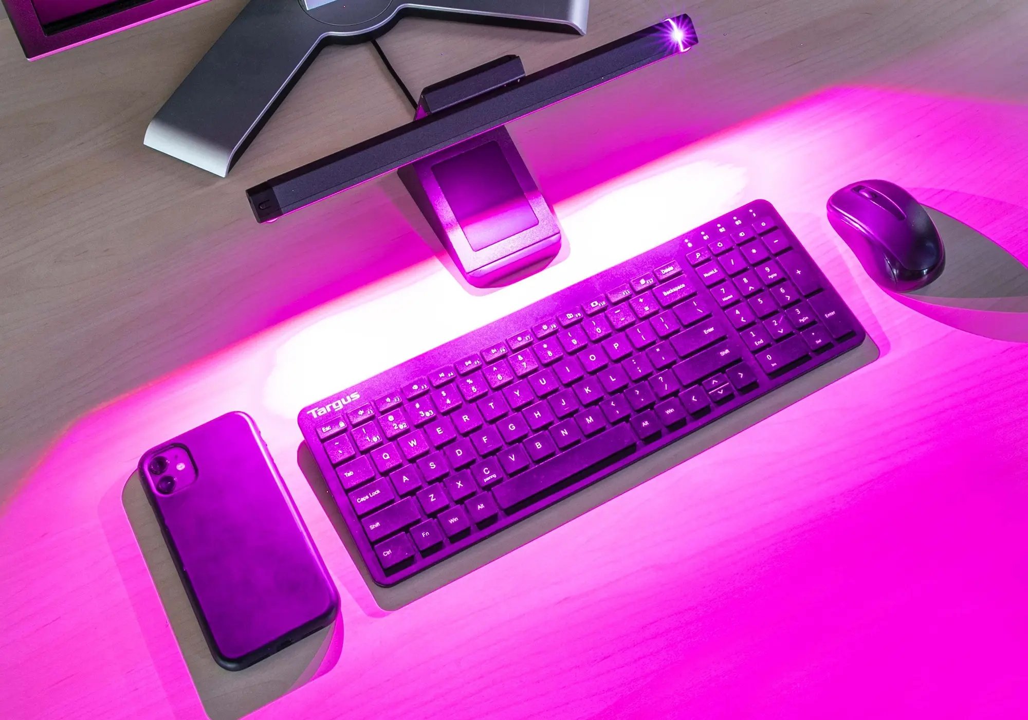 A workstation setup with a keyboard, mouse, smartphone, and monitor illuminated by bright pink lighting from a desk-mounted light bar.