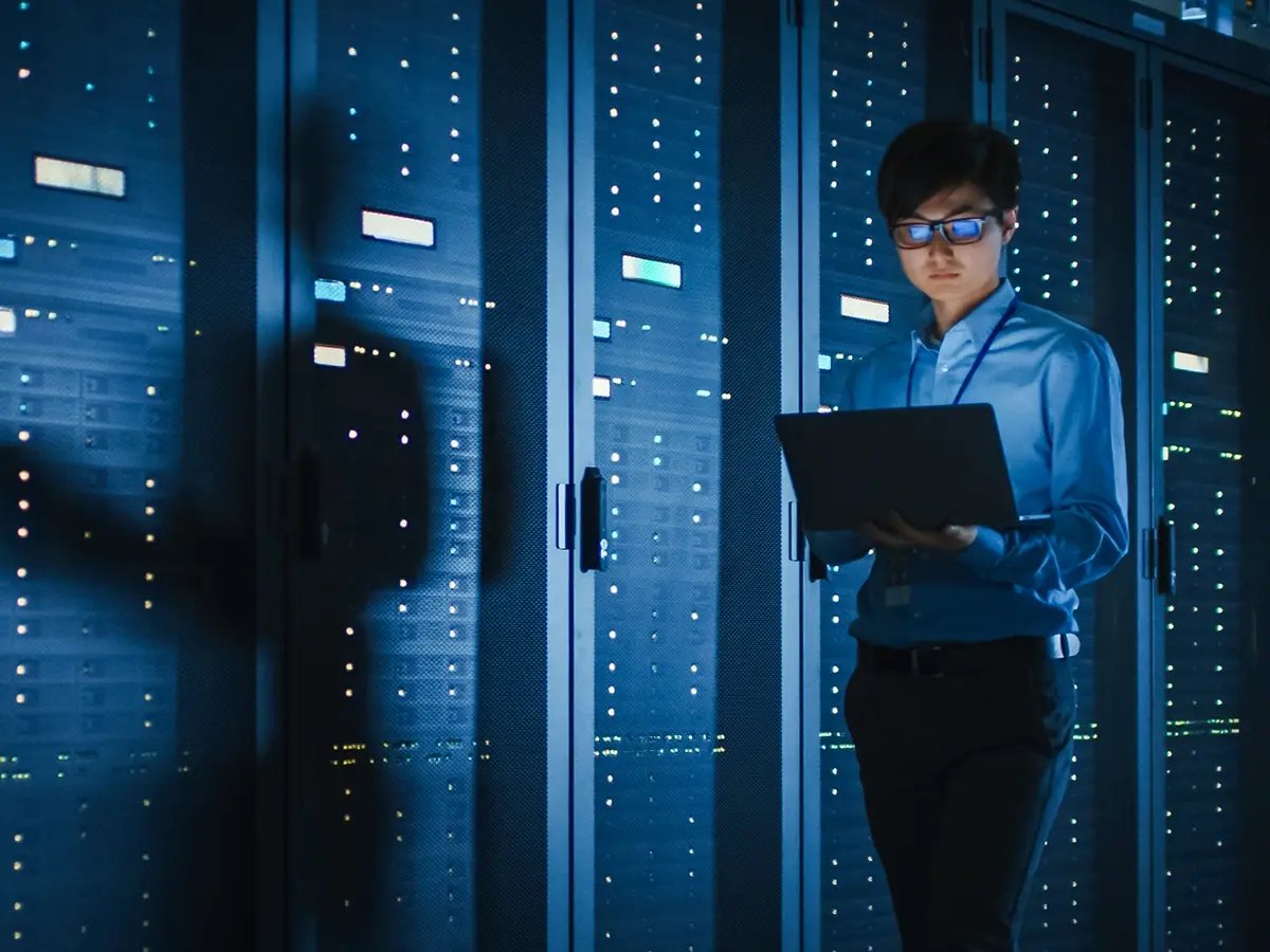 Person standing in a server room holding a laptop, surrounded by rows of illuminated server racks with blue indicator lights.