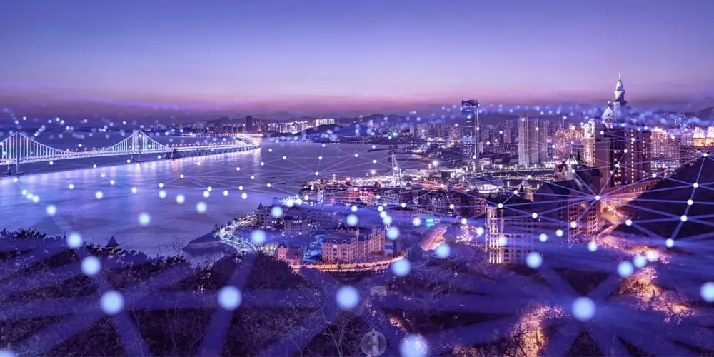 City skyline at dusk with illuminated buildings and a bridge over water, overlaid with glowing blue network connection lines representing digital connectivity.