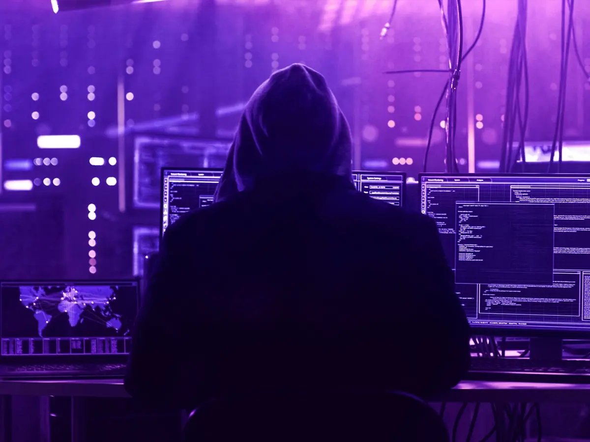 Person wearing a hoodie seated at a desk with multiple monitors displaying code and data, in a dark room illuminated by purple light, representing cybersecurity threats.