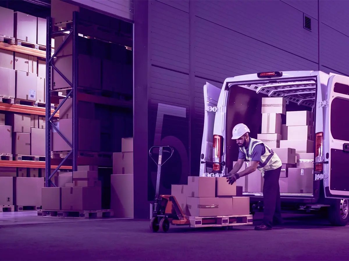 Worker loading cardboard boxes onto a pallet jack next to a van filled with packages inside a warehouse with shelves stacked with more boxes.