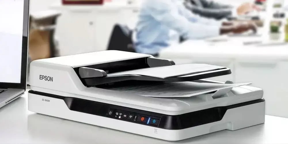 Epson scanner with documents being scanned, placed on a desk next to a laptop in an office setting.
