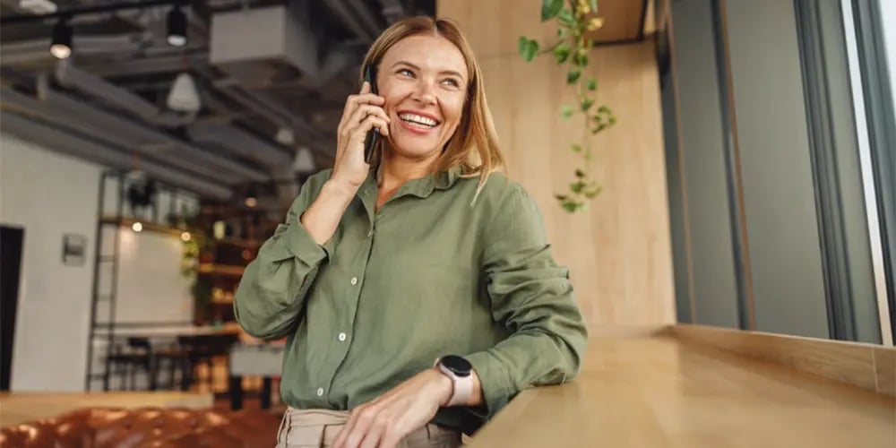 A person wearing a green long-sleeve shirt is standing by a wooden counter near large windows, holding a smartphone to their ear in a modern indoor setting with plants and industrial-style decor.