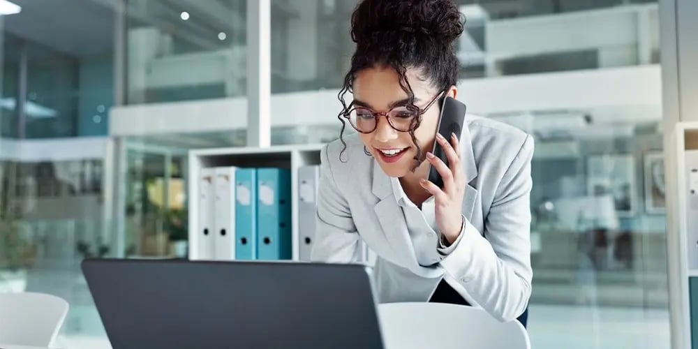 Person in a light-colored blazer is leaning over a desk, talking on a smartphone while working on a laptop in a modern office setting.
