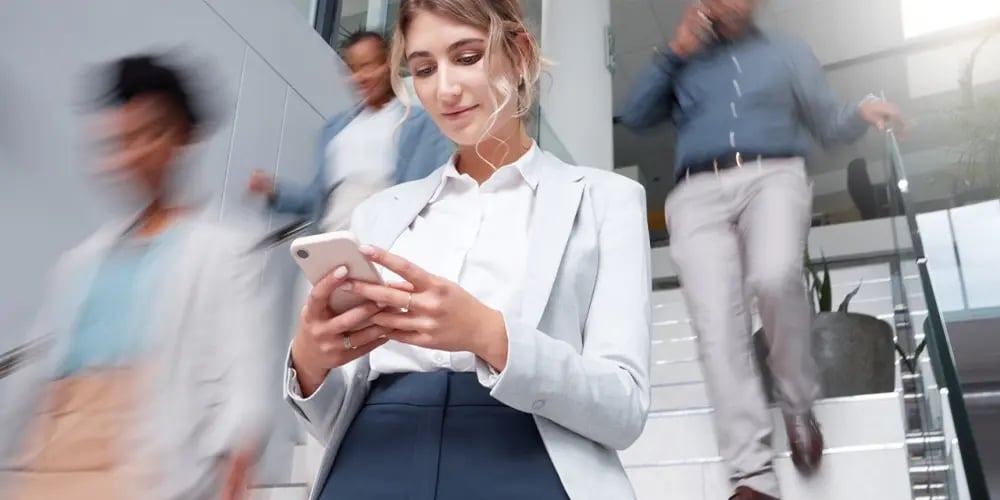 Person in business attire is standing on a staircase, looking at a smartphone, with others walking down the stairs in the background.