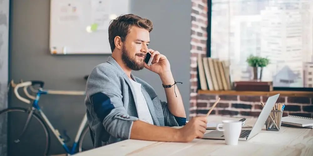 Person sitting at a desk in a modern office, talking on a phone while using a laptop, with a bicycle and bookshelf in the background.