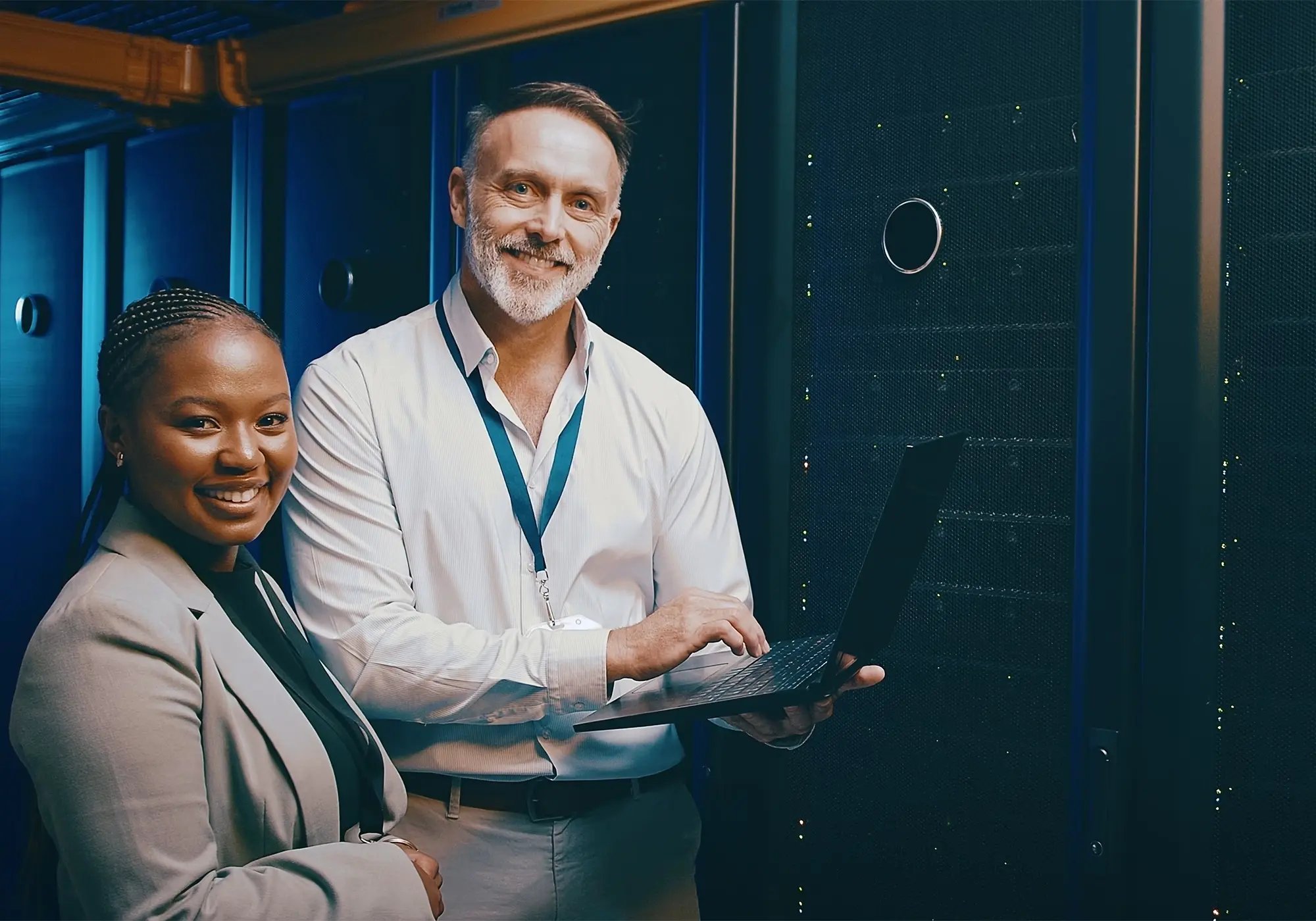Two people in business attire stand in a server room, one holding a laptop while discussing.